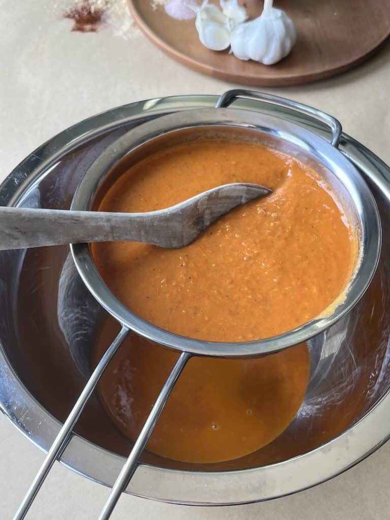 tomato and onion sauces in a metal strainer over a metal bowl with wooden spoon to make a sauce for Beef Enchiladas with Flour Tortillas