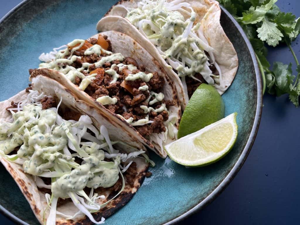 ground beef tacos with avocado cilantro aioli sauce on flour tortillas with lime wedges