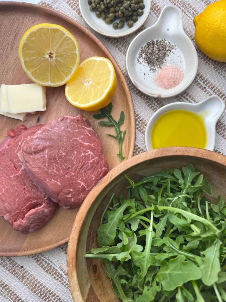 top sirloin steak, lemons, butter, capers, salt and pepper, oil, arugula in bowls and on a plate