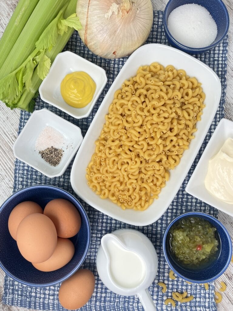 dried macaroni pasta, salt and pepper, mustard, relish, sugar, mayo and eggs in bowls, with milk in a pitcher. Celery and onion on the table